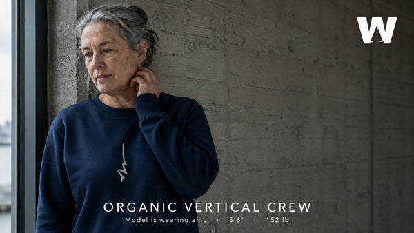 A woman leaning against a concrete wall by a window, wearing the French Navy Organic Vertical Crew sweatshirt.