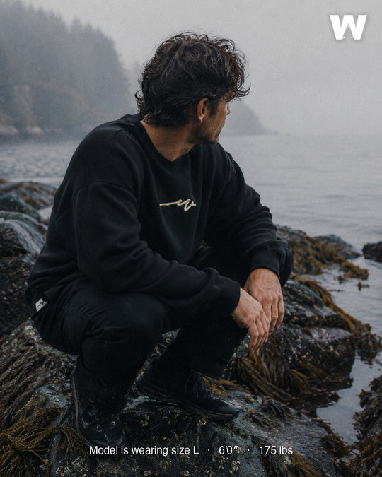 Model wearing the Black Puff Dub Heavy Terry Crew crouched on a foggy Salish Sea shoreline — size L