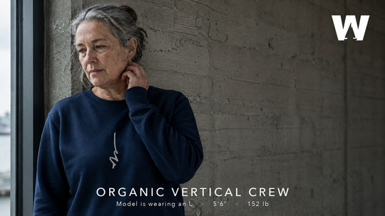 A woman leaning against a concrete wall by a window, wearing the French Navy Organic Vertical Crew sweatshirt.