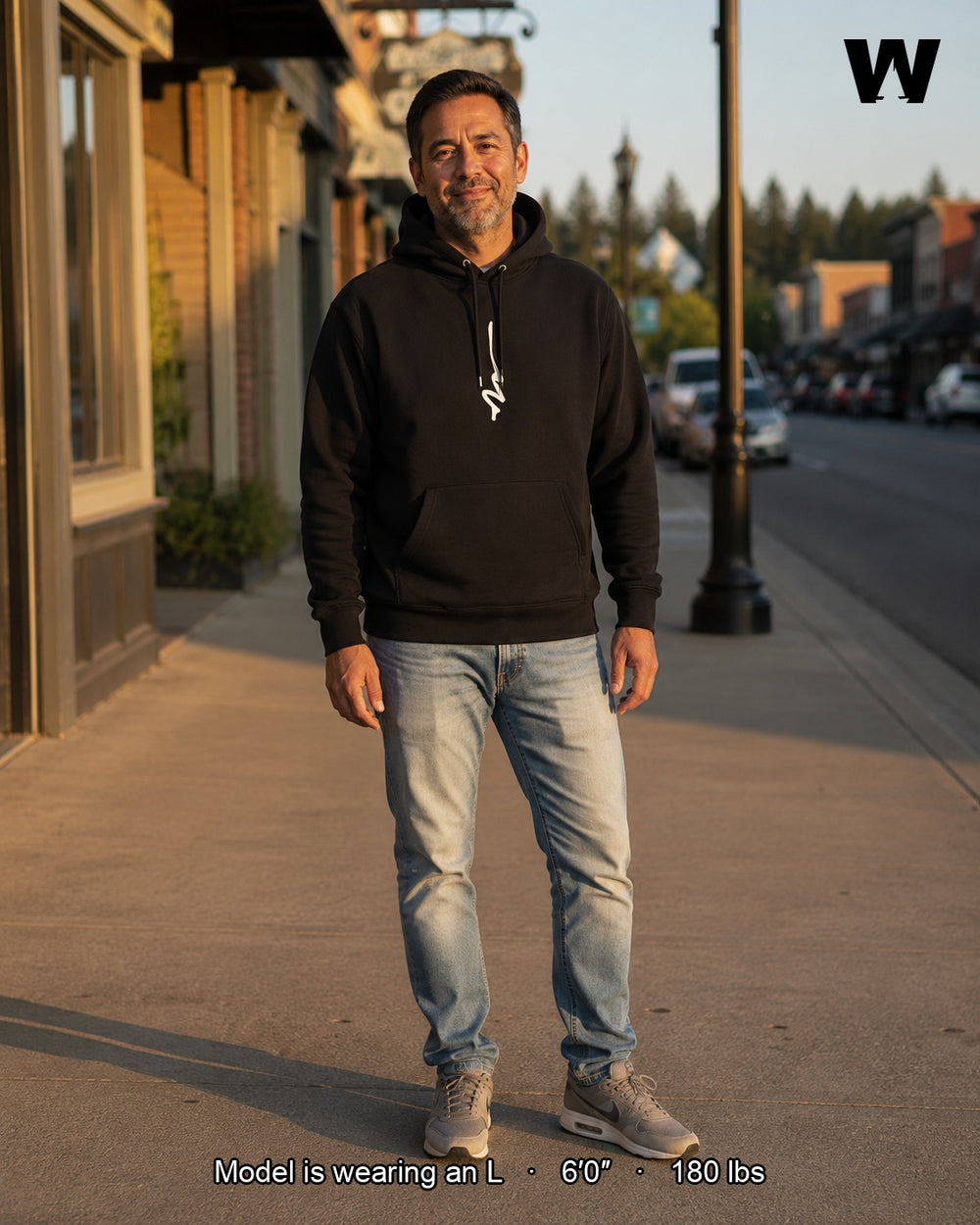 A man of mixed Caucasian and Hispanic heritage stands on a First Street sidewalk in Snohomish, Washington at golden hour wearing the black Silky Vertical Hoodie (size L) with light-blue jeans and grey sneakers.