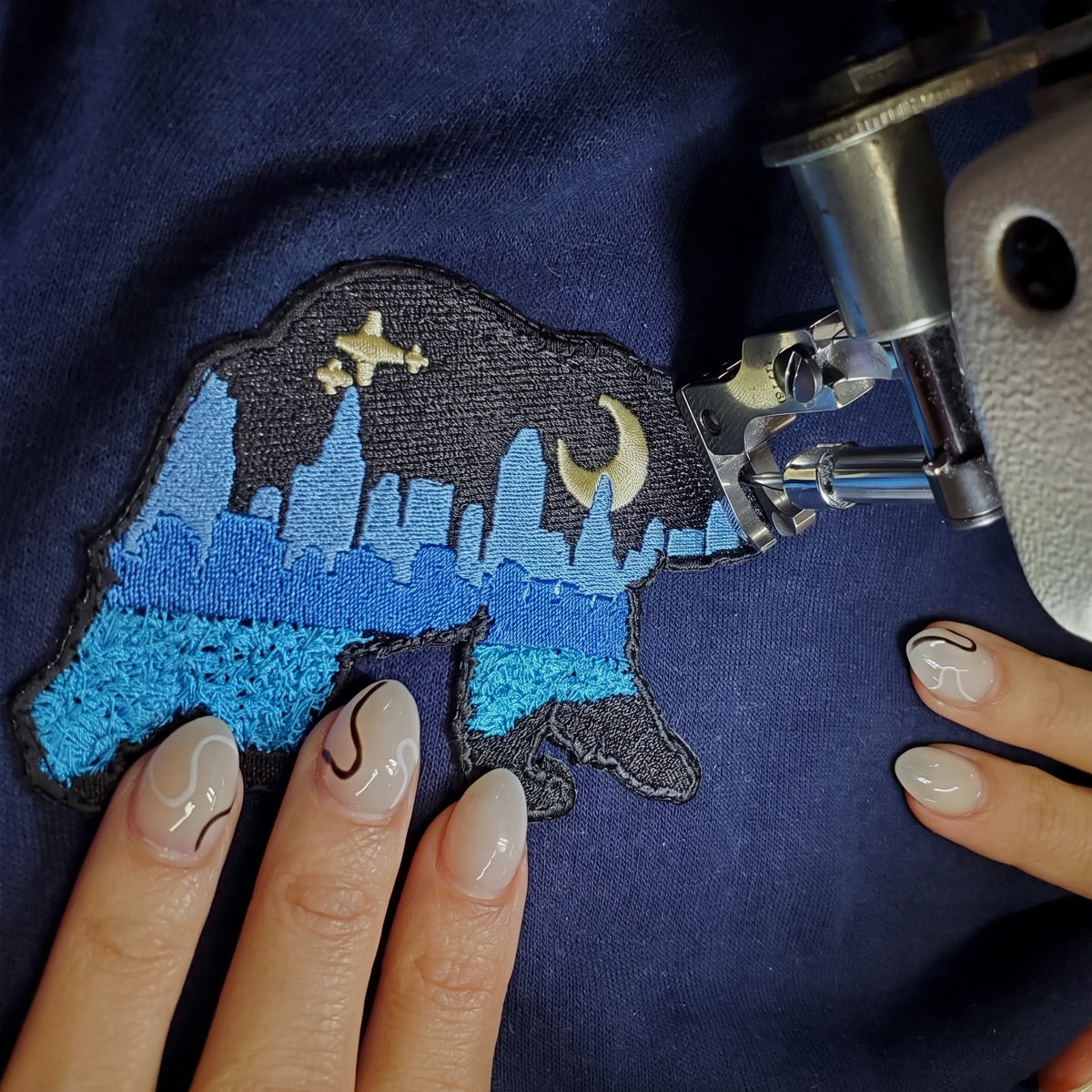 A seamstress guides the At The City patch under a sewing machine as it is stitched onto a navy garment.